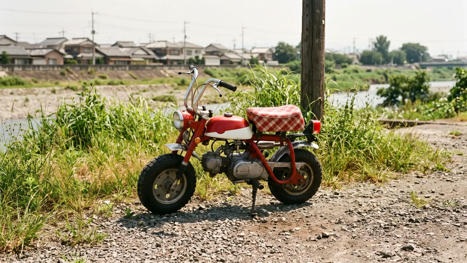 昭和42年〜昭和46年(1967〜1971年ごろ)のホンダのモンキーを再現したイメージ。空き地の砂利の上に止めた、小さな太いタイヤと短い車体、チェック柄の座席を持つ赤と白の小型のバイク。