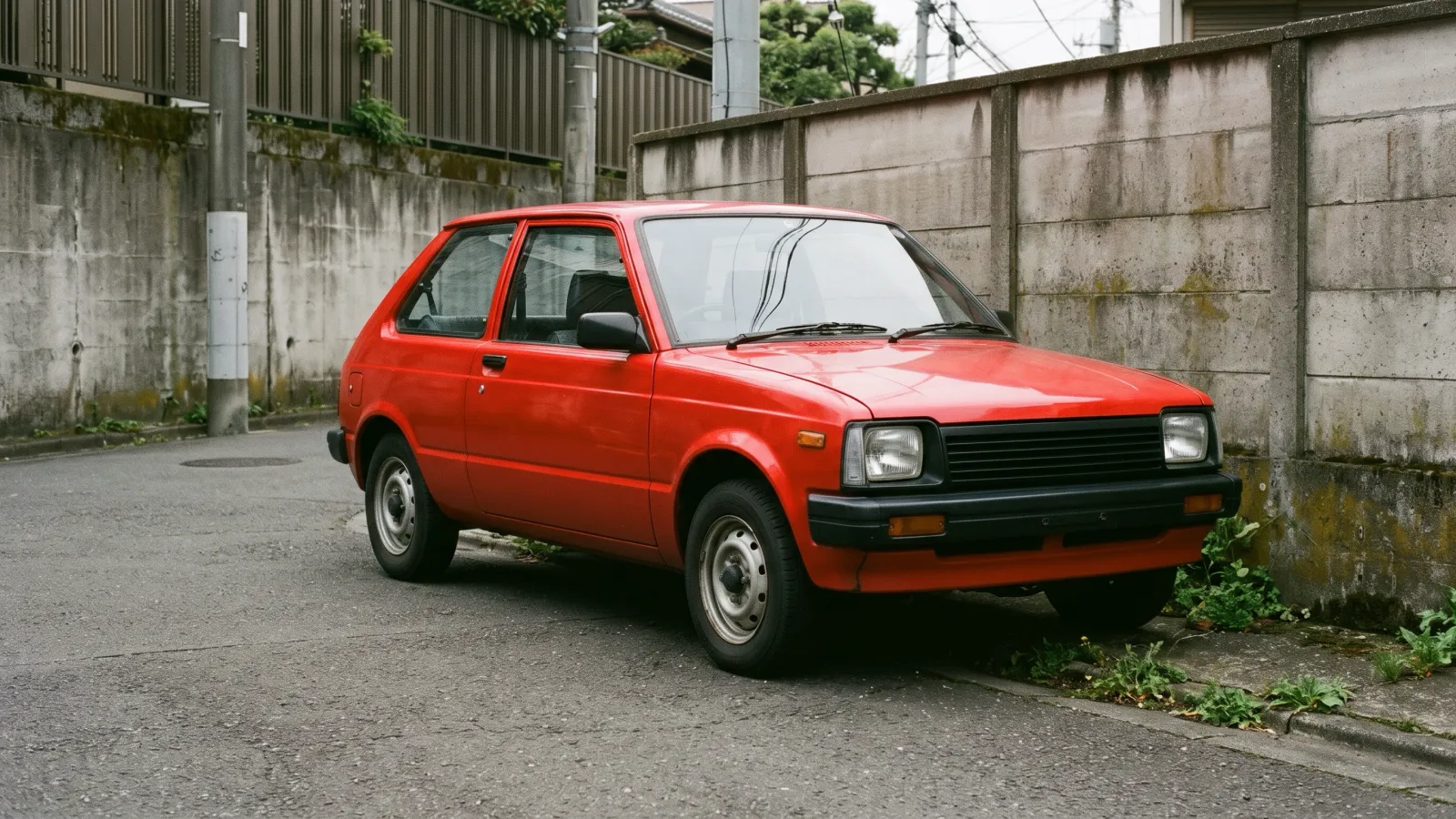 昭和57年〜昭和61年（1982〜1986年ごろ）の日産のマーチを再現したイメージ。低いコンクリート塀の脇の静かな住宅街に、四角いヘッドライトと黒い格子を持つ赤い角ばった三ドアの小型車が春の光の中で止まっている。